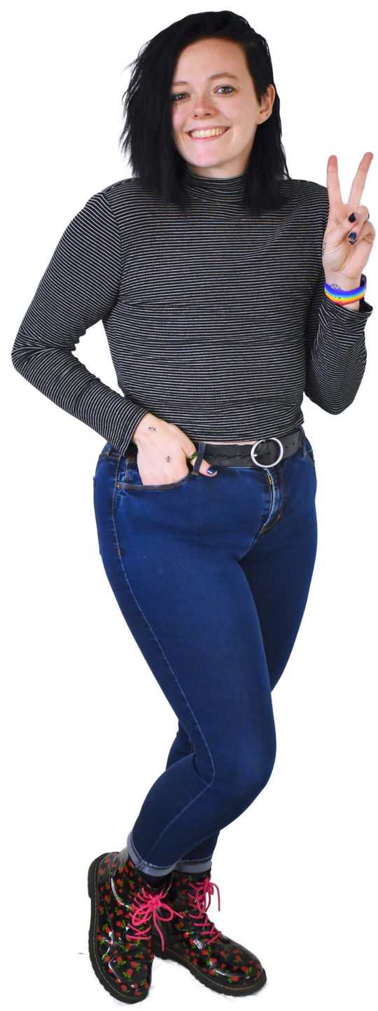 A full-body photo of Charlie, who's wearing a striped long-sleeve shirt, a rainbow watch, jeans with a belt, and colorful boots. They're smiling and holding up a peace sign.