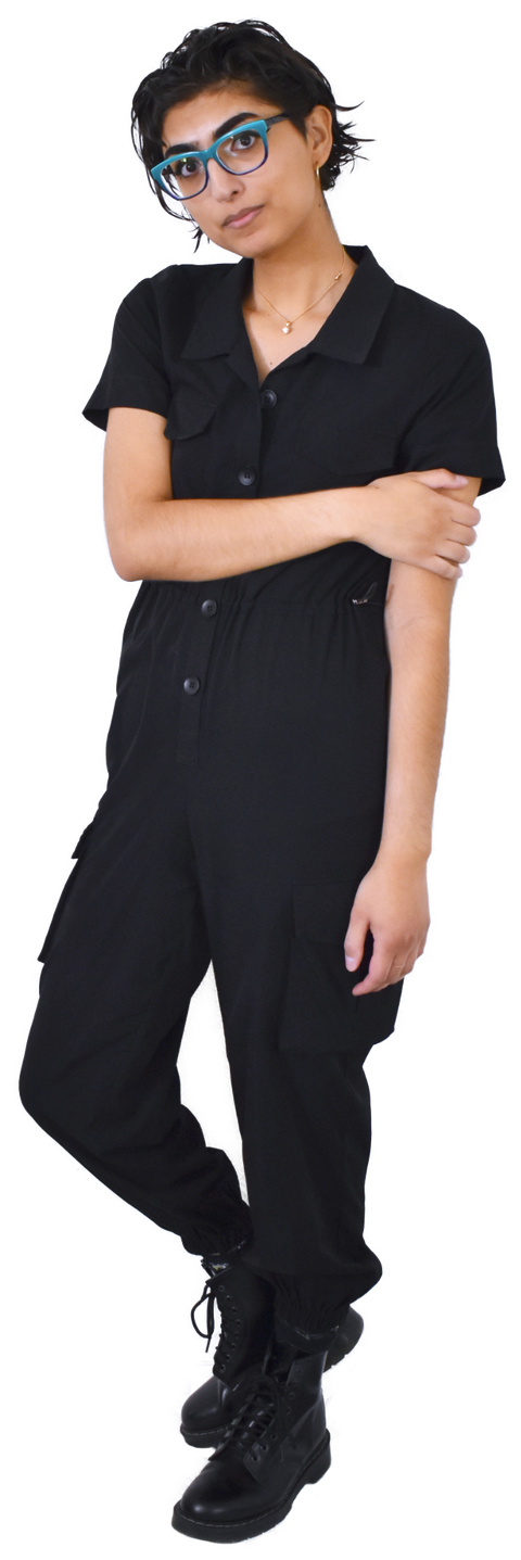 A full-body photo of Stacy, who is wearing a simple necklace with a black jumpsuit and black boots. They're holding their arm.