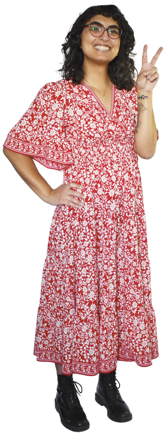 A full-body photo of Daisy, who is wearing a simple necklace with a red and white floral dress and black boots. She's doing a peace sign.